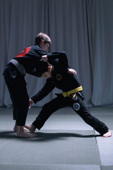 Two young boys engage in jiu jitsu practice wearing gis on a mat indoors.